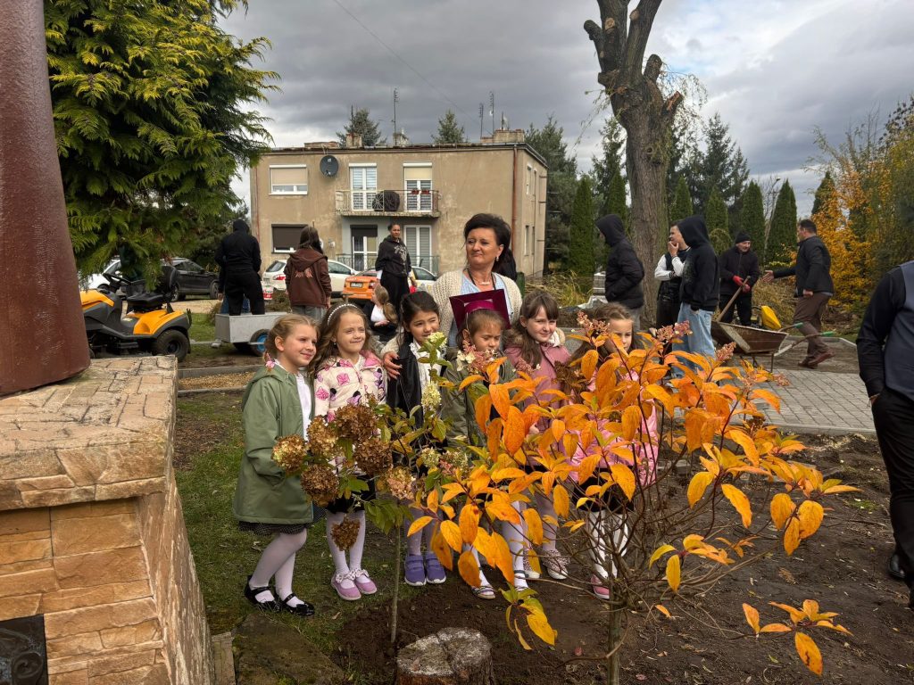 Dzieci podczas uroczystości pasowania na ucznia na zewnątrz, w jesiennej scenerii, z nauczycielem w tle.