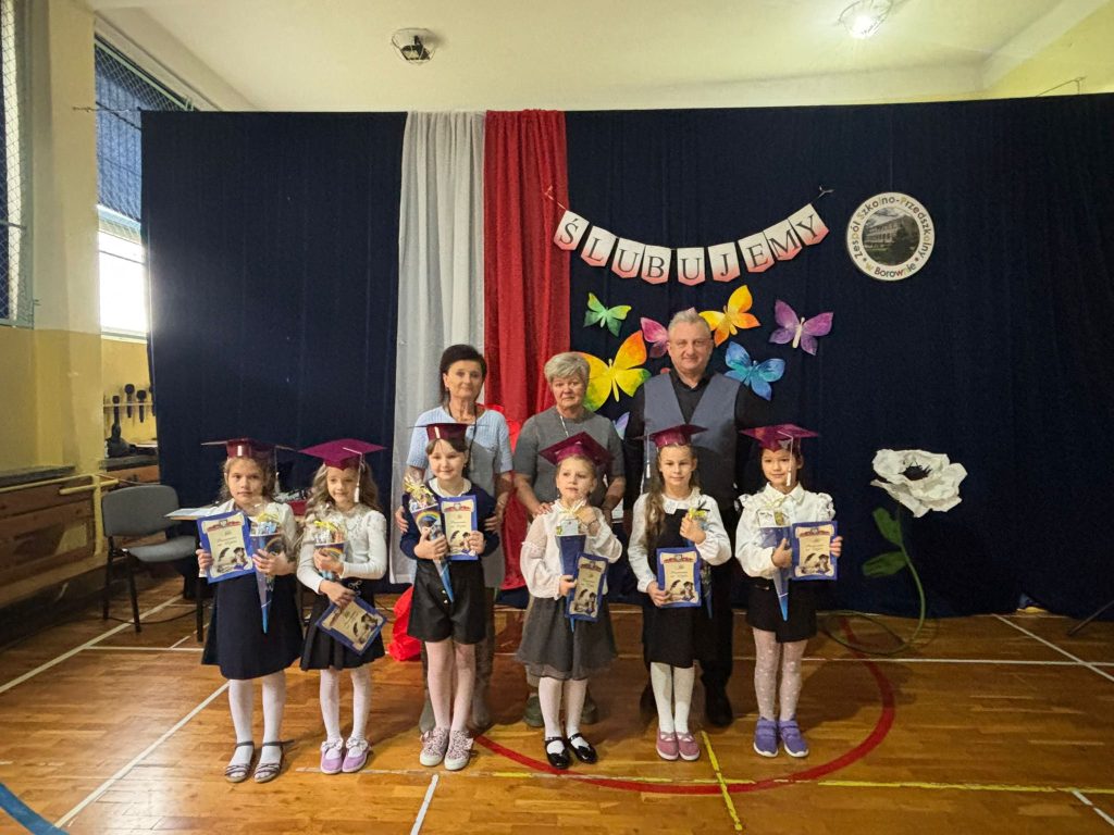 Uczniowie w biretach z dyplomami i upominkami podczas pasowania na ucznia na tle szkolnej dekoracji.