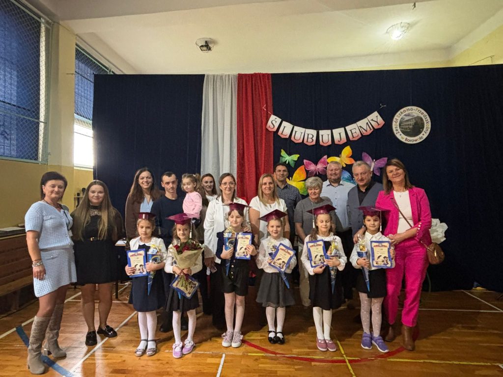 Fragment uroczystości pasowania na ucznia: poczet sztandarowy oraz dzieci ustawione przed sceną w sali gimnastycznej.