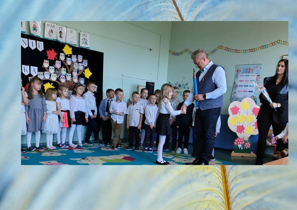 Uroczystość pasowania w klasie: dyrektor wręcza pierwszoklasistce symboliczny duży ołówek, a reszta dzieci stoi w szeregu w odświętnych strojach.