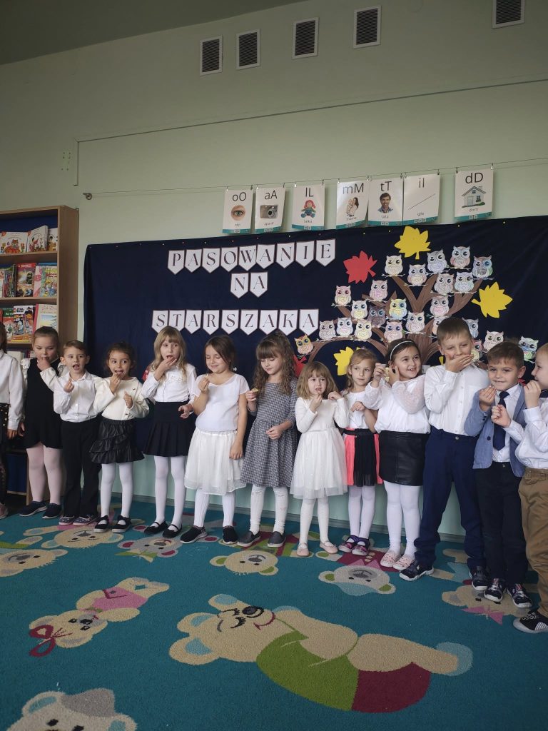 Grupa dzieci w odświętnych strojach stoi przed dekoracją z napisem „Pasowanie na starszaka” i drzewem z papierowymi sowami.