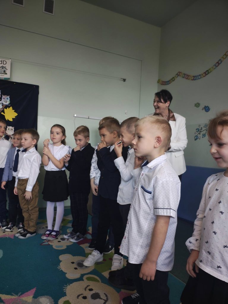 Dzieci stoją podczas uroczystości pasowania w klasie; w tle uśmiechnięta nauczycielka obserwuje występ.