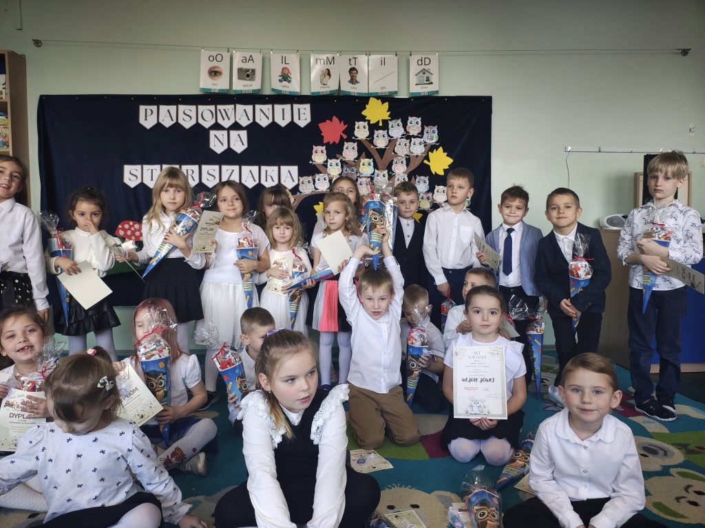 Grupa dzieci z dyplomami i upominkami pozuje w klasie przed dekoracją „Pasowanie na starszaka”; w tle papierowe sowy na drzewie.