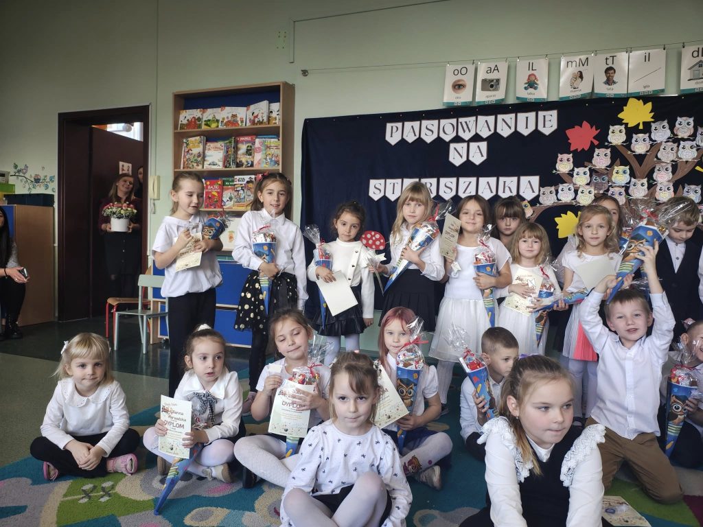 Zdjęcie grupowe dzieci z dyplomami i upominkami na tle dekoracji „Pasowanie na starszaka”; część dzieci siedzi na dywanie, część stoi.