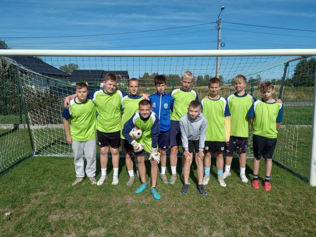 Grupa młodych piłkarzy pozujących do wspólnego zdjęcia na boisku piłkarskim. Chłopcy stoją przed bramką, ubrani w zielone stroje sportowe, jeden z nich trzyma piłkę i ma na sobie rękawice bramkarskie.