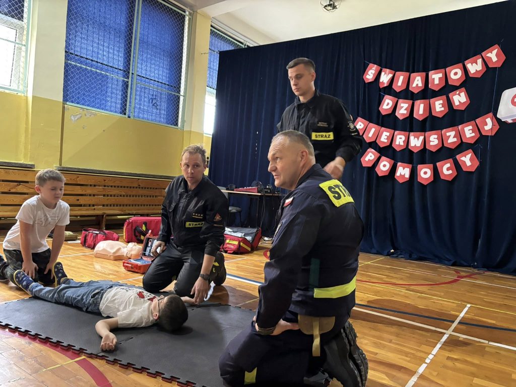 Strażacy prowadzą zajęcia z pierwszej pomocy dla dzieci w sali gimnastycznej. Dziecko leży na macie, a obok znajduje się sprzęt ratowniczy i torba medyczna. W tle dekoracja z napisem Światowy Dzień Pierwszej Pomocy.
