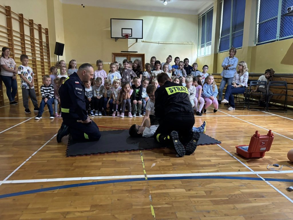 Pokaz udzielania pierwszej pomocy w sali gimnastycznej. Strażacy demonstrują czynności ratunkowe na leżącym dziecku, a grupa dzieci siedzi i obserwuje wydarzenie.