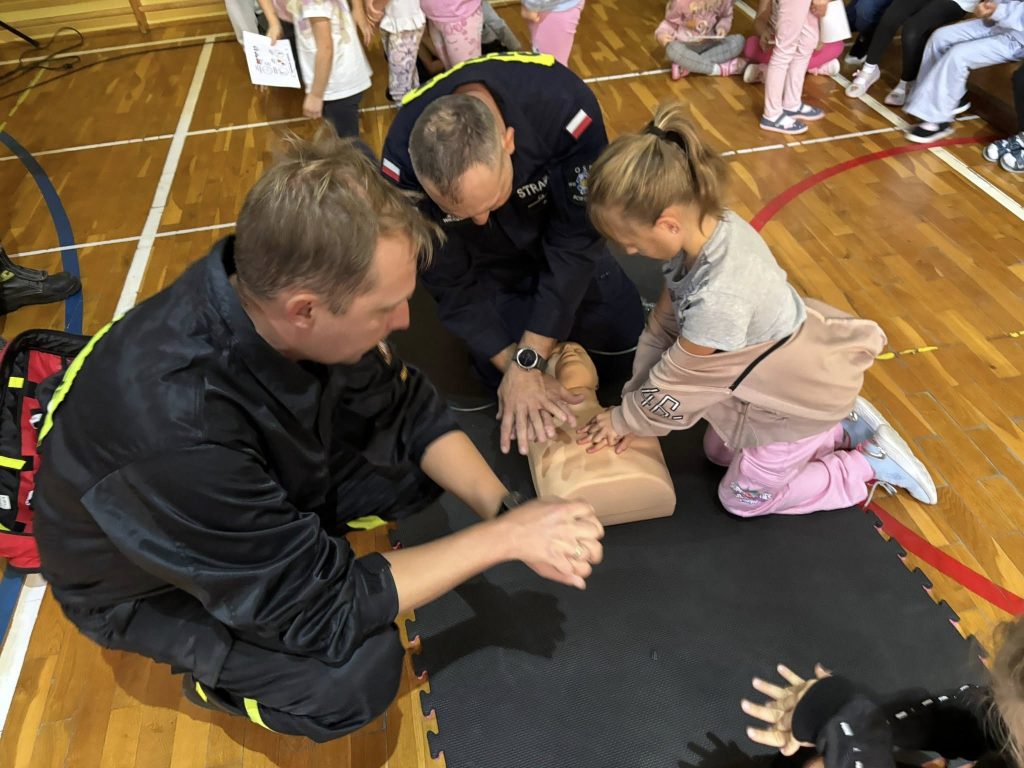 Strażacy prowadzą pokaz pierwszej pomocy w sali gimnastycznej. Dziecko wykonuje uciski klatki piersiowej na fantomie pod nadzorem strażaków, wokół stoją inne dzieci.