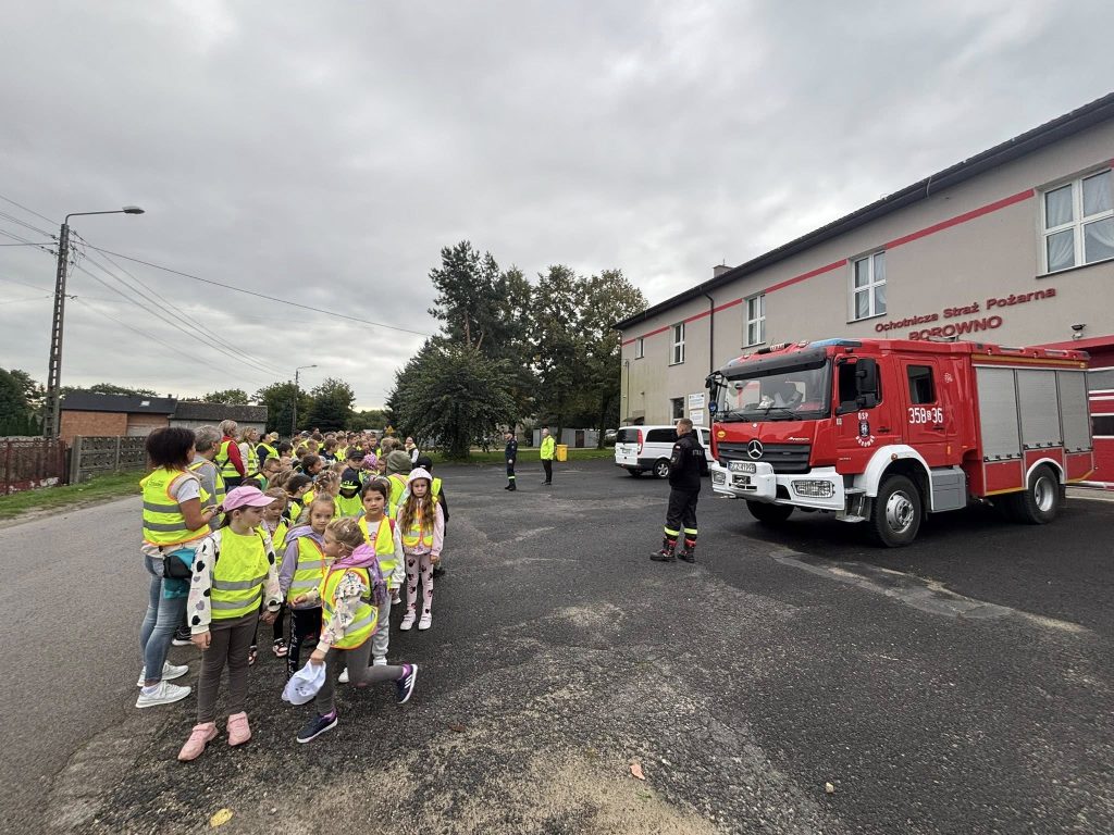 Grupa dzieci w kamizelkach odblaskowych stoi na placu przed budynkiem straży pożarnej. Obok zaparkowany jest czerwony wóz strażacki, a strażak porządkuje ustawienie uczestników.