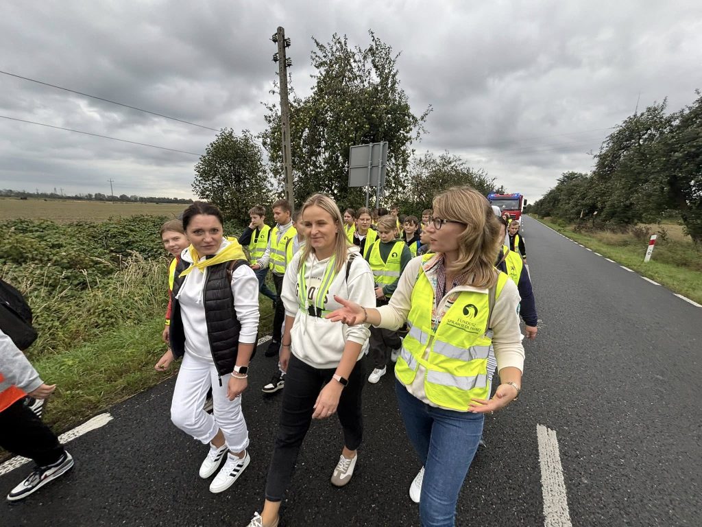 Grupa dzieci i opiekunów w kamizelkach odblaskowych idzie poboczem drogi poza terenem zabudowanym. Na czele grupy idą osoby dorosłe prowadzące kolumnę pieszych.