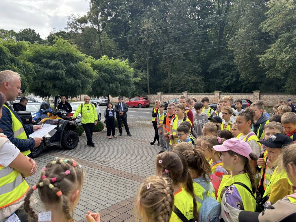 Grupa dzieci w kamizelkach odblaskowych stoi na placu przed budynkiem straży pożarnej. Obok zaparkowany jest czerwony wóz strażacki, a strażak porządkuje ustawienie uczestników.