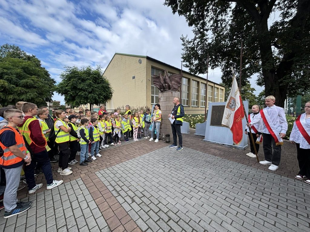 Uczniowie w kamizelkach odblaskowych stoją w skupieniu przed pomnikiem. Obok znajduje się poczet flagowy z biało-czerwoną flagą, a osoba dorosła prowadzi wystąpienie.
