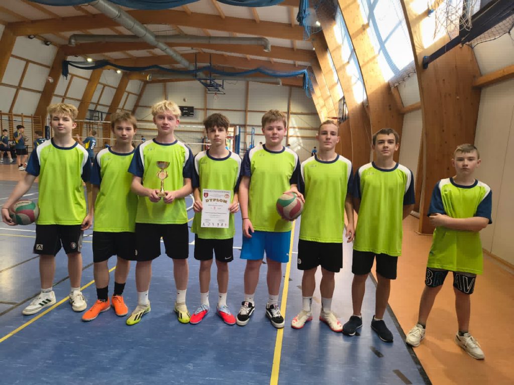 Grupa młodych siatkarzy w zielonych koszulkach stoi w hali sportowej. Dwóch chłopców trzyma puchar i dyplom, dwóch ma piłki.