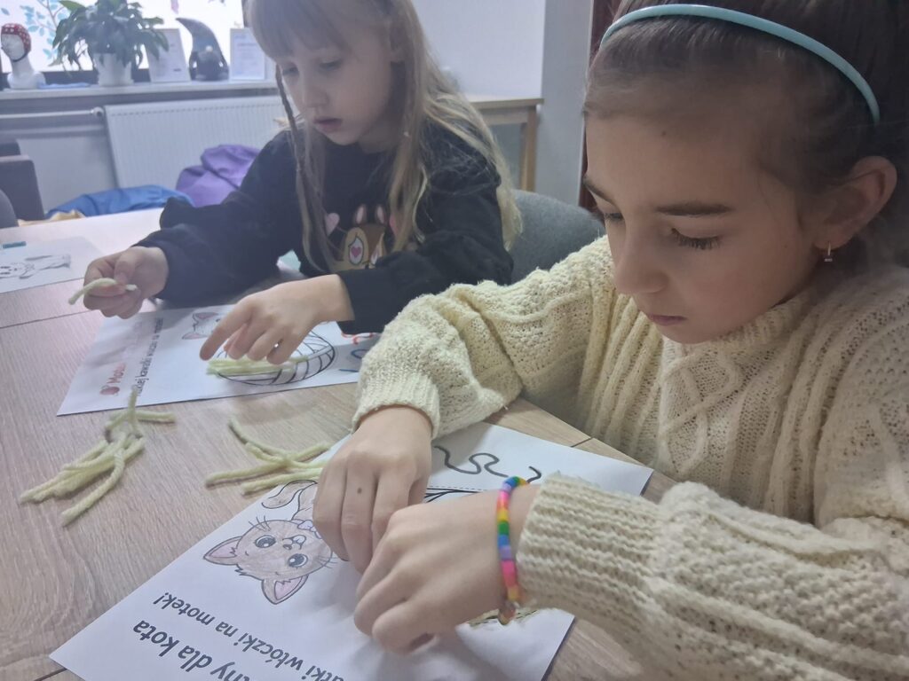 Dwie dziewczynki przy stole przyklejają kawałki włóczki do kartki z rysunkiem kota.