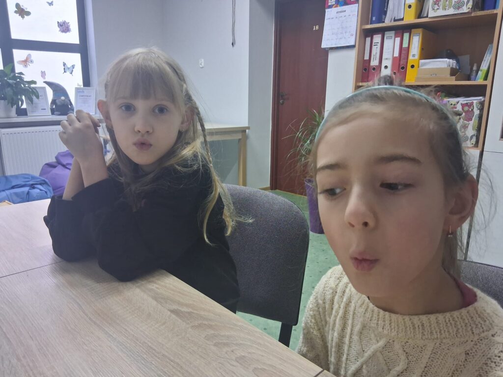 Dwie dziewczynki siedzą przy stole i wykonują ćwiczenie logopedyczne z układaniem ust w kształt „O”.