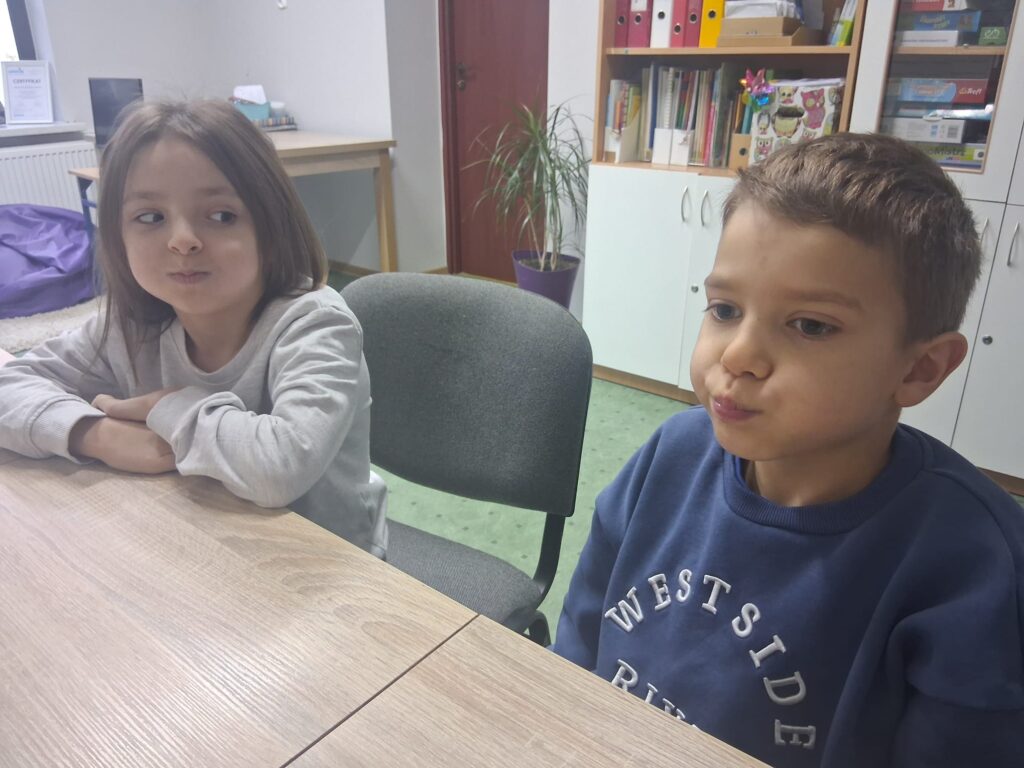 Dziewczynka i chłopiec siedzą przy stole i wykonują ćwiczenie logopedyczne z nadymaniem policzków.