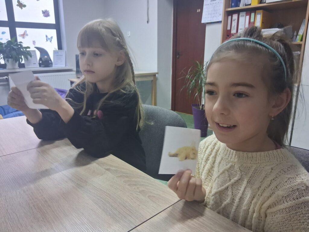 Dwie dziewczynki siedzą przy stole i pokazują małe kartki z obrazkami.