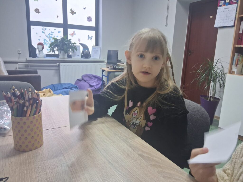 Dziewczynka siedząca przy stole pokazuje kartki z obrazkami podczas zajęć.