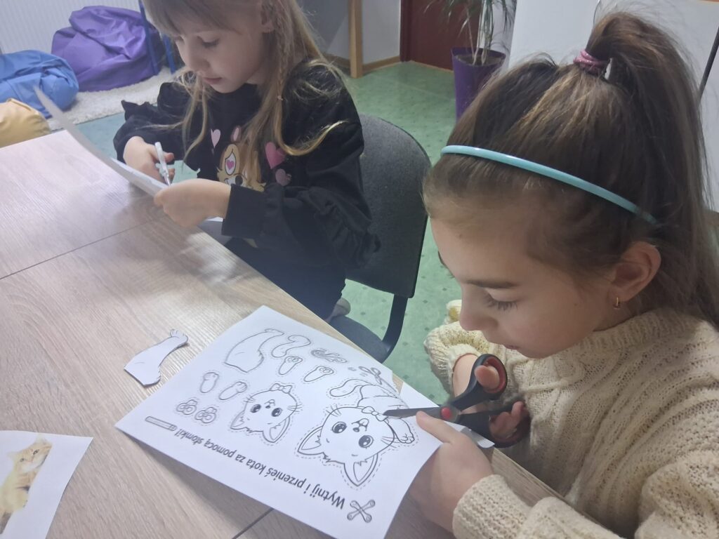 Dwie dziewczynki siedzą przy stole i wycinają elementy z kartki z rysunkiem kota podczas zajęć plastycznych.