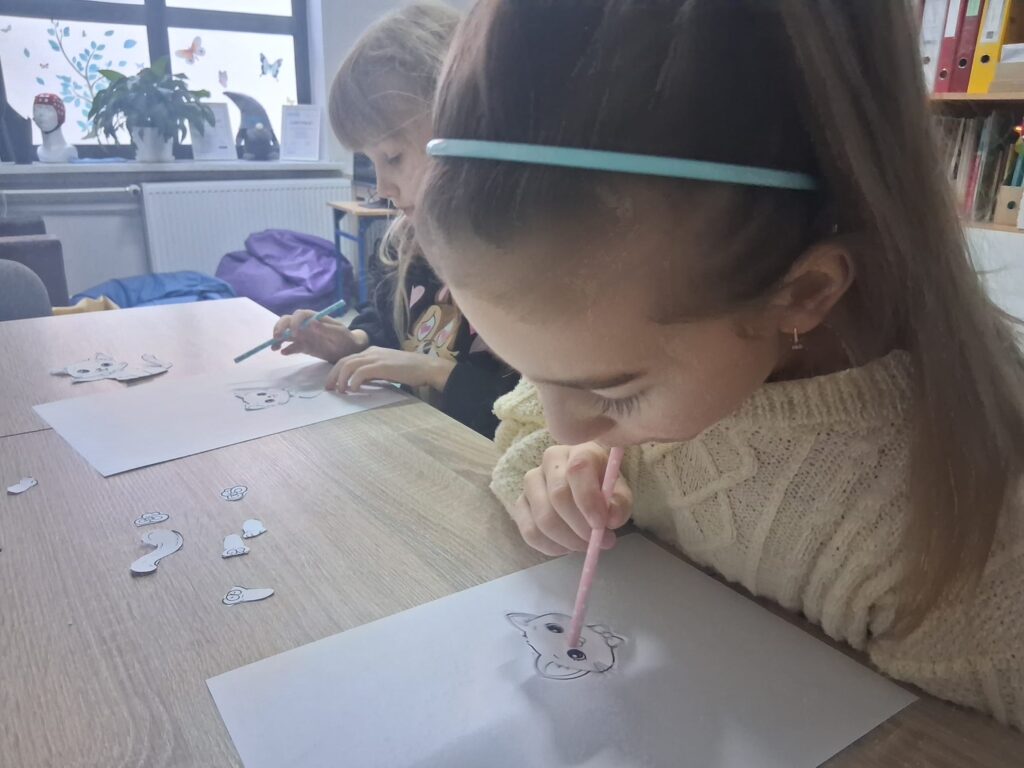 Dziewczynka koloruje rysunek kota na kartce przy użyciu kredki, a druga dziewczynka rysuje obok przy stole.