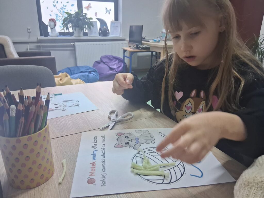 Dziewczynka wykonuje pracę plastyczną z włóczki na kartce z rysunkiem kota i kłębka wełny.