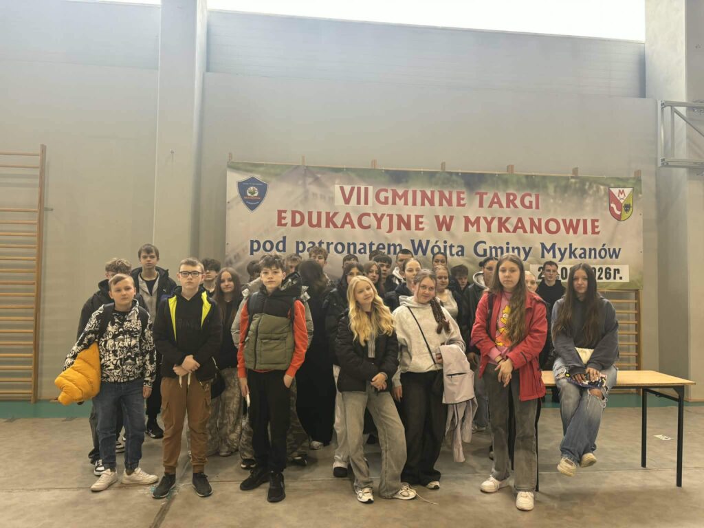 Grupa uczniów pozuje w hali sportowej na tle baneru „VII Gminne Targi Edukacyjne w Mykanowie”, prezentując swoją szkołę podczas wydarzenia.